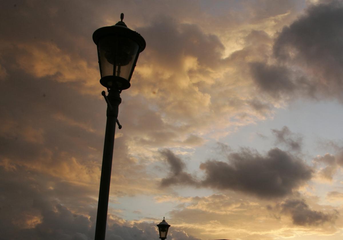 La silueta de dos farolas se recortan en un atardecer entre nubes y claros.