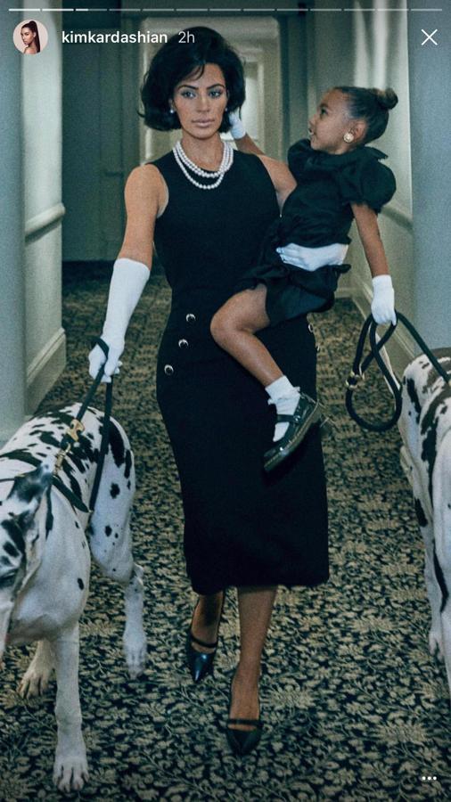 Kim Kardashian en el pasillo de un edificio con un vestido negro largo, cogiendo su hija en brazos y llevando dos perros atados.