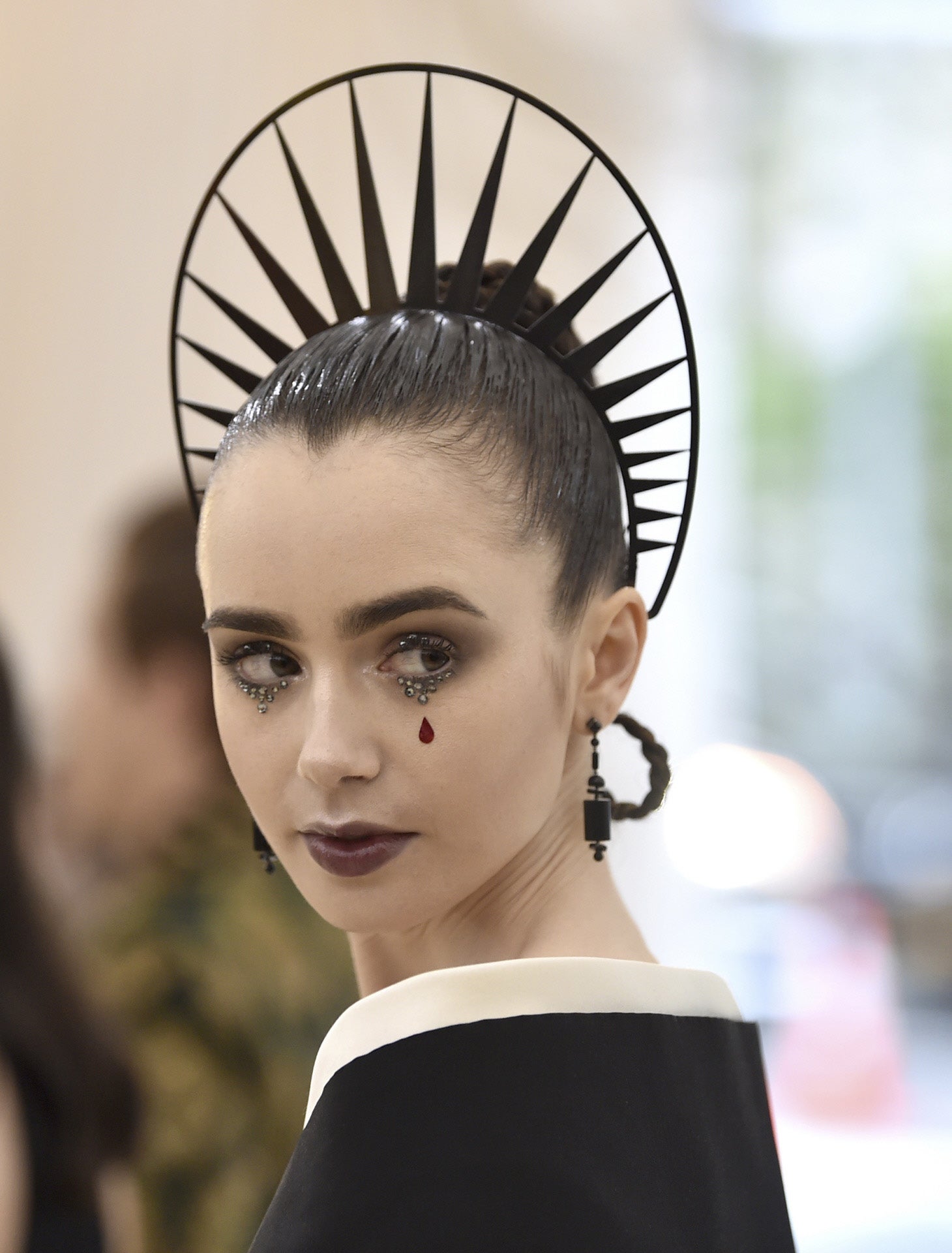 Lily Collins con moño de trenzas efecto mojado y adornado con una corona negra. ¡Ojo al maquillaje! La lágrima de sangre y las perlitas son lo más y su color de labios muy acertado.