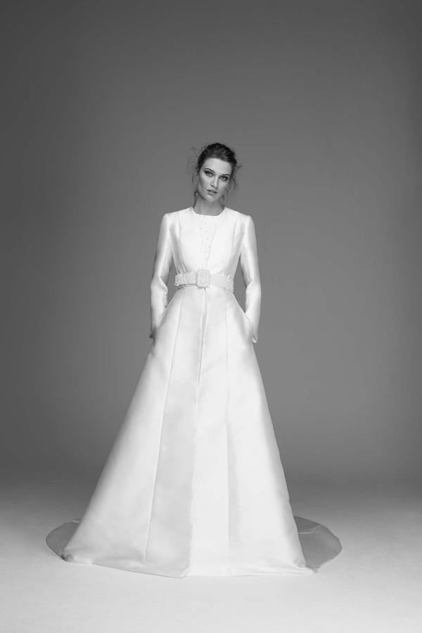 Vestido de crepé velado con tul de perlas bordadas, con escote a la caja y sin mangas. Encima, abrigo de mikado con manga sastre y escote pico, que se cierra con un cinturón de perlas.