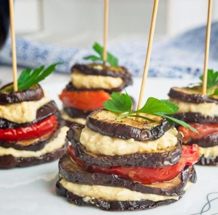 Ingredientes (para 5 unidades):1 berenjenaHummus2 tomates asados o tomates deshidratados en aceiteSalAOVEPreparación:Lava y seca la berenjena. Corta en rodajas finas de medio centímetro. Echa sal y coloca en un plato. Mete al microondas durante 5 minutos para que se cocinen ligeramente.Pon una plancha o sartén a calentar. Echa un poco de aceite en la plancha y dora las rodajas de berenjena por ambos lados.Separa varios montones de las rodajas de berenjena por tamaños. De esta manera podrás ir colocando de mayor a menor tamaño. Necesitas 4 rodajas por cada uno de estos montaditos.Unta la rodaja más grande de humus generosamente. Coloca encima la siguiente rodaja de berenjena. Ahora pon el tomate asado. Coloca otra rodaja de berenjena más pequeña y unta también con humus. Pon la última rodaja de berenjena, en esta ocasión la más pequeña de todas y pincha todas las capas con un palillo. Y ya tienes listo otro aperitivo para triunfar.