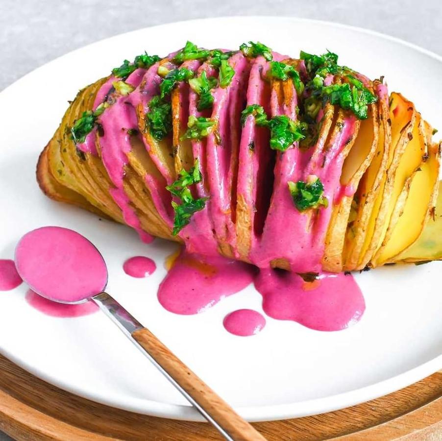 Ingredientes:2 patatas grandes1 yogur de soja50 gr de remolacha cocidaAjoPerejil frescoAOVEPimienta negra molidaSalPreparación:- Precalentar el horno a 200°C.- Lavar bien las patatas.- Realizar cortes transversales que no lleguen hasta la base de la patata, para ello te puedes a ayudar de un palillo largo tipo brocheta con el que se atraviesa la base de la patata y hace de tope para el cuchillo.- Disponer las patatas en una bandeja de horno y añadir un chorrito de aceite, sal y pimienta negra bien distribuido entre los cortes.- Hornear 1 h aprox. (dependerá del tamaño de las patatas y del horno). A los 30 min impregnarlas con una mezcla de ajo y perejil picados. Hornear otros 30 min. o hasta que estén completamente asadas.- Para la salsa: Triturar el yogur y la remolacha junto con un chorrito de aceite, sal y pimienta.- Para servir bañar la patata con la salsa y añadir un poco más de la picada de ajo y perejil por encima.