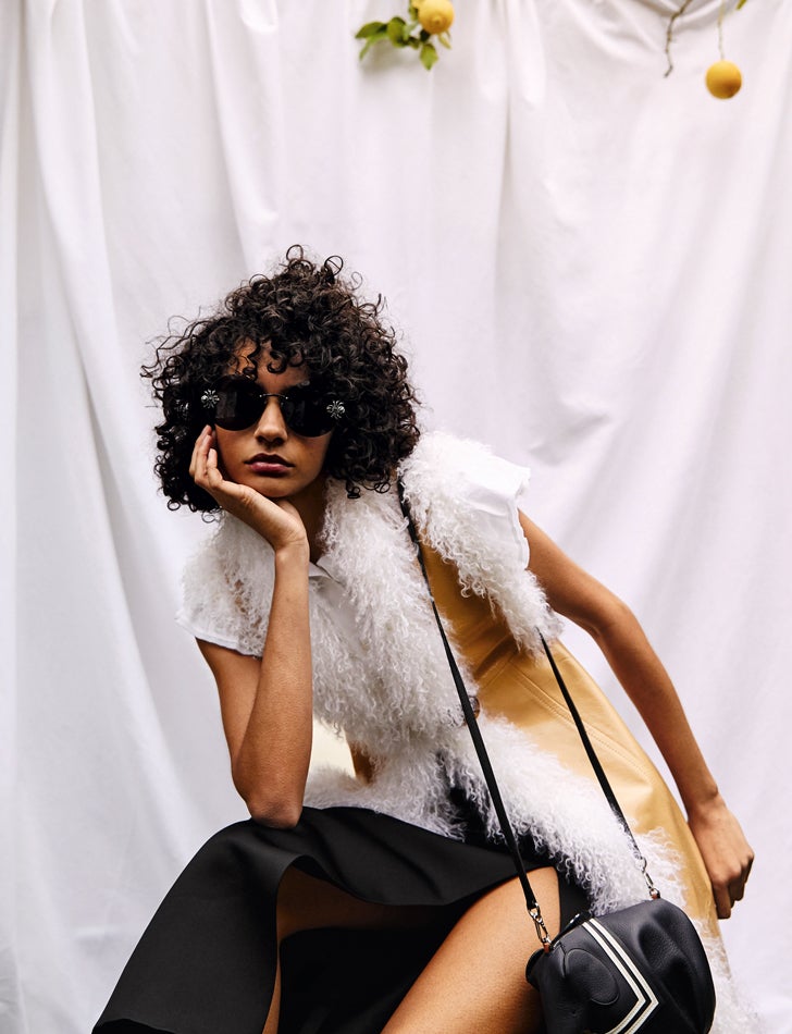 Chaleco largo de cuero y lana rizada, blusa de algodón de manga corta y falda, todo de Miu Miu. Las gafas de sol son de Alexander McQueen y el bolso Elefante Sailor, de Loewe.