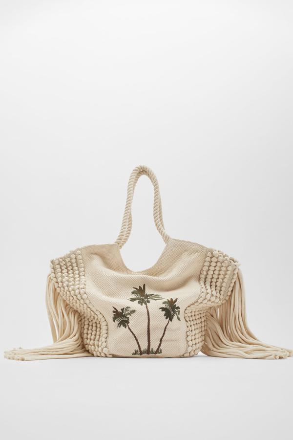 Este bolso en formato shopper de color beige con detalle de palmeras bordadas en la parte delantera cuenta con laterales trenzados con detalles de flecos. Con asas de hombro de cuerda, tiene un precio de 39,95 euros.