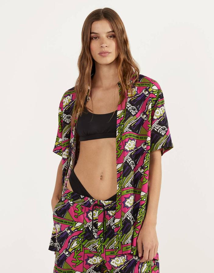 Fondo rosado con estampado en motivos verdes y negros con botellas de Coca-Cola para esta camisa de Bershka con mangas cortas que tenía  un precio de 22,99 euros  y está disponible ahora por 9,99 euros entre las tallas XS y XL.