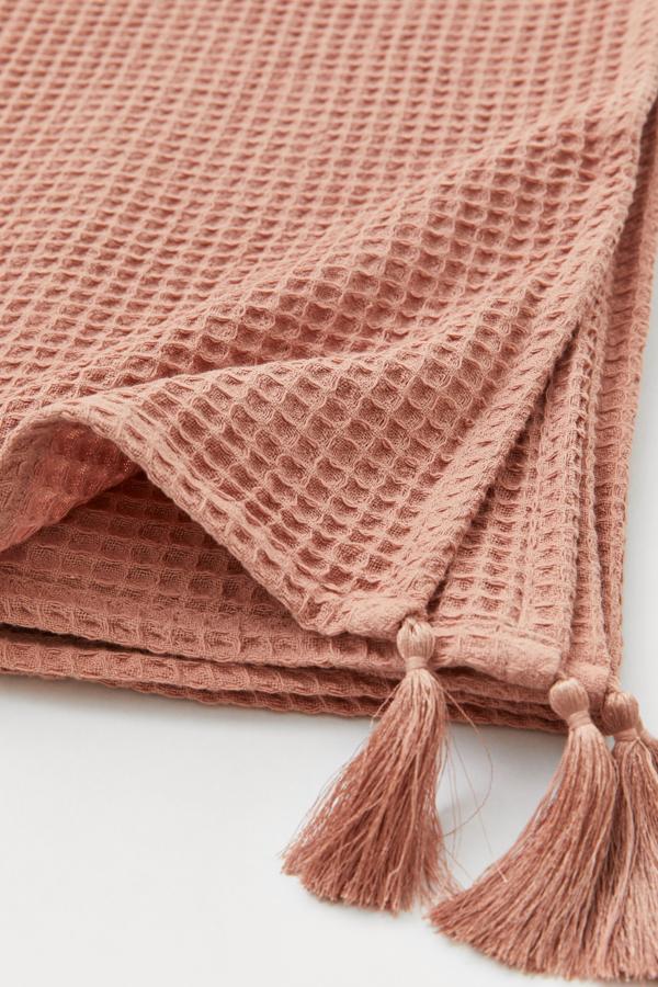 En color rosa palo, y disponible en otros colores, esta manta en tejido gofrado de algodón con borlas en las esquinas mide 100x150 centímetros y tiene un precio de 14,99 euros.