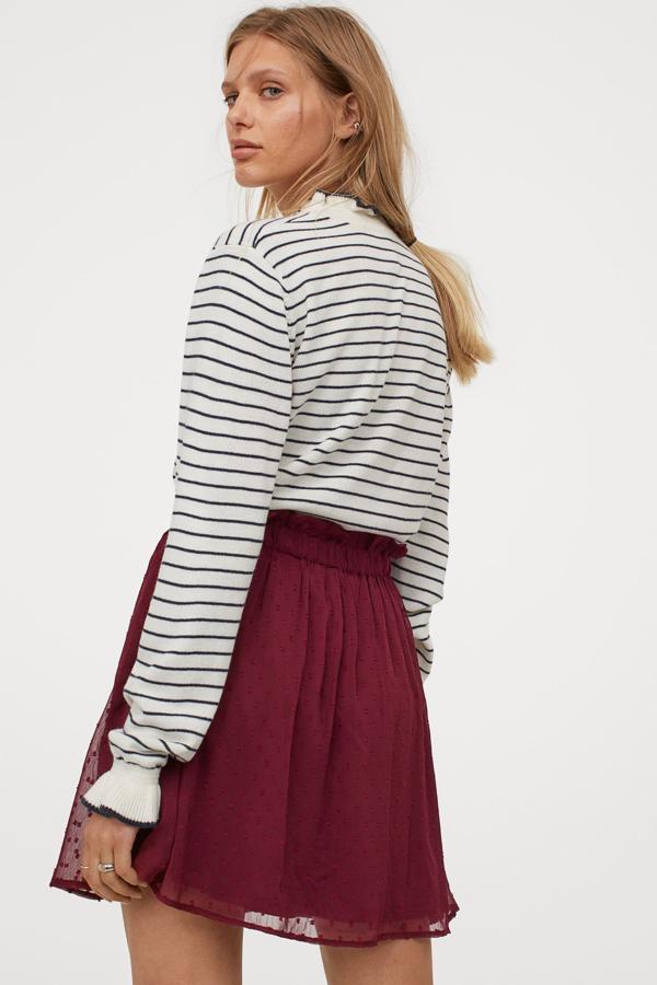 En color rojo oscuro, esta falda corta en gasa de plumeti con vuelo cuenta con ribete de volante en la cintura y tiene un precio de 19,99 euros. Está disponible en otros colores.