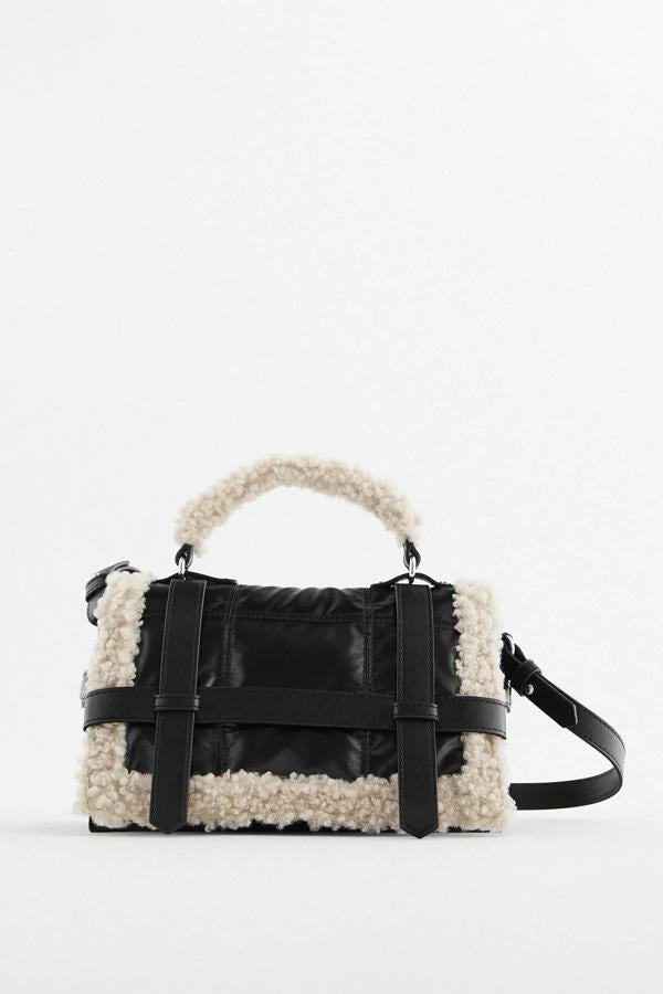 En color negro con contorno en tejido efecto borreguillo, este bolso formato city con solapa acolchada con detalle de tiras cuenta tanto con asa de mano como de hombro. Tiene un precio de 35,95 euros.