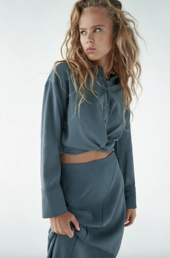 Con cuello de solapa y cierre frontal con botones, esta camisa fluida de Zara en color plomo viene con bajo con nudo en delantero y elástico en la espalda. Está disponible entre las tallas XS y XXL, menos en la L, por 22,95 euros.