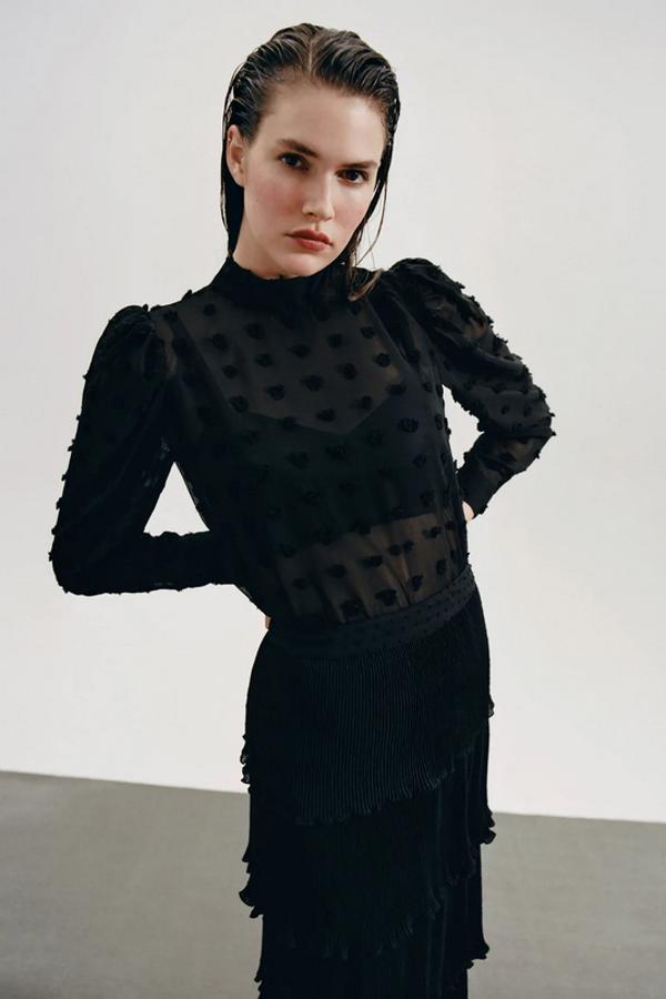 De cuello subido y manga larga con hombreras, esta blusa semitransparente en plumeti de Zara viene con cierre en la espalda. Cuesta 25,95 euros y está disponible entre las tallas XS y XL.