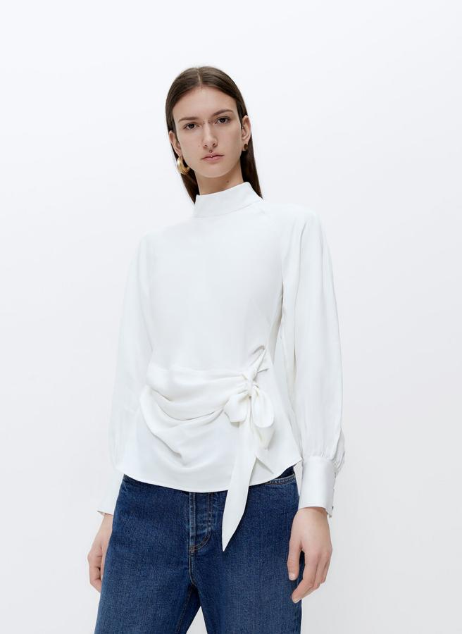 En color crudo, confeccionada en viscosa de alta calidad, cuello perkins con abertura de cremallera y manga raglán larga abullonada, esta camisa viene con detalle de drapeado con lazada en delantero y está disponible en las tallas S, M y L. De 79 euros ha pasado a costar 49,95 euros.