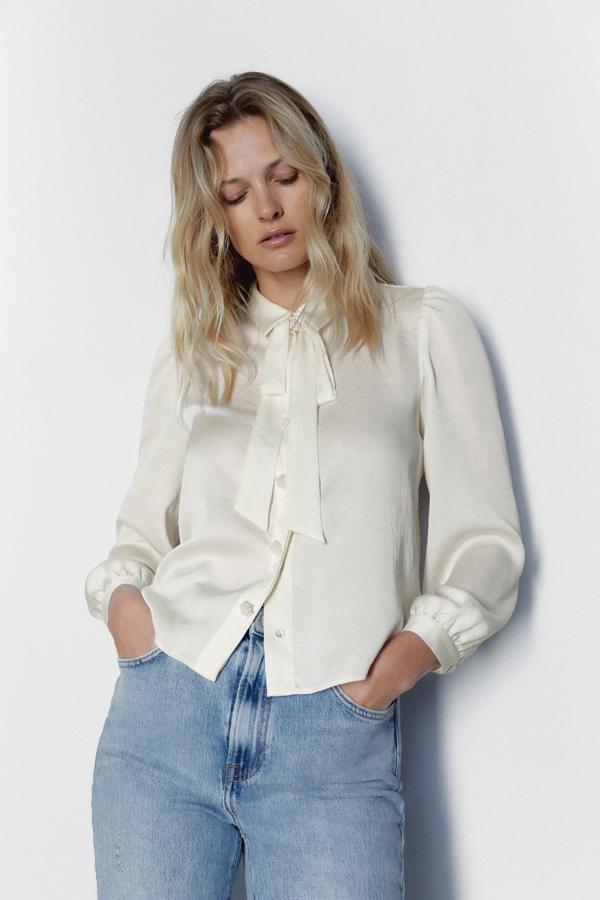 Esta blusa de satén blanco roto con lazo al cuello y botones joya será el complemento perfecto de tus vaqueros, pero también de tus trajes, faldas midi e incluso looks de invitada de primavera. Cuesta 25.95 euros en Zara.