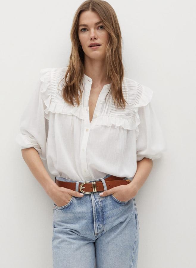 En color blanco roto, esta blusa fluida en tejido semitransparente cuenta con cuello mao y detalles de volantes y plisados. Tiene un precio de 29,99 euros.