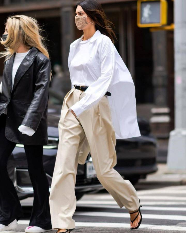La supermodelo lleva camisa 'oversize' y camiseta blanca junto a pantalón con pinzas en beige, que ajusta al cuerpo con un cinturón. Resuelve el look con sandalias destalonadas en negro.