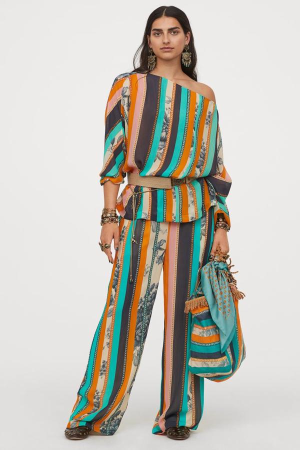 Este pantalón largo en crepé de gasa con suave caída y estampado de rayas verticales en tonos marino, esmeralda y naranja es de talle alto y viene con elástico revestido en la cintura y cordón de ajuste. Cuesta 39,99 euros. La blusa a juego es vaporosa y ligeramente arrugada, de corte cuadrado y escote amplio que deja un hombro al descubierto. Tiene un precio de 34,99 euros. 