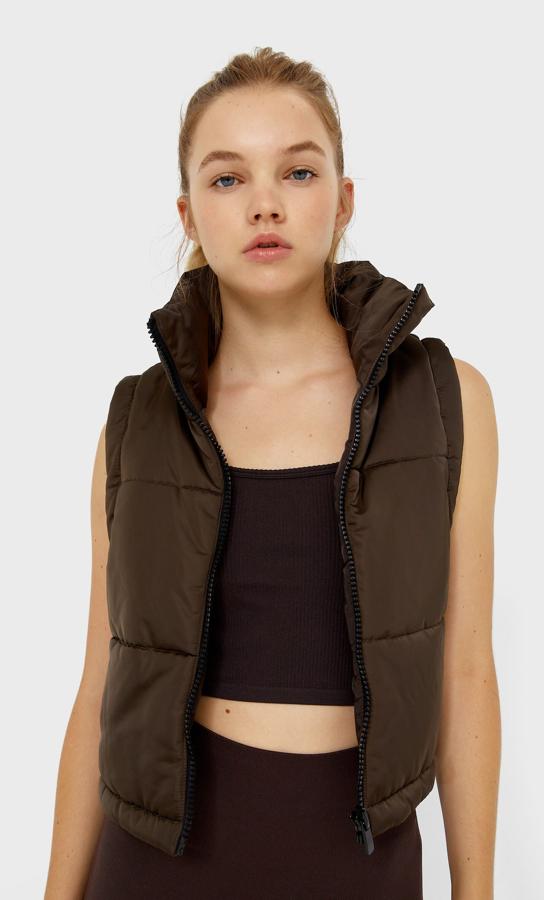 En color marrón y tejido satinado, este chaleco cropped de Stradivarius con cuello subido y manga sisa tiene cierre de cremallera frontal y cuesta 25,99 euros. 