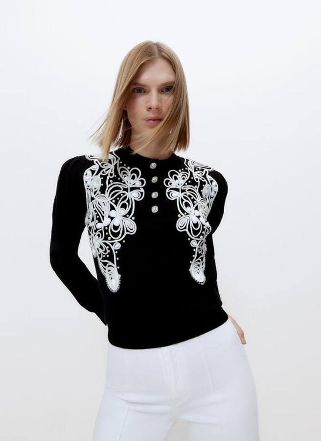 En color negro, con detalle de bordado y pedrería a contraste en blanco, este jersey de punto con cuello redondo abierto con botones joya viene con mangas largas con frunces y acabados en rib. Tiene un precio de 99 euros y está disponible en las tallas S y M.