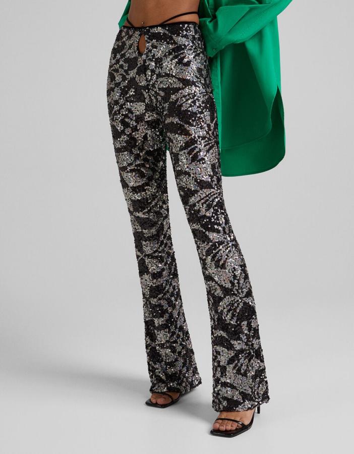 Con tiras y cut out en la cintura, este pantalón de lentejuelas de Bershka en plateado y negro es de diseño flare con bajos ligeramente acampanados y cuesta 39,99 euros. Está disponible entre las tallas XS y L. 