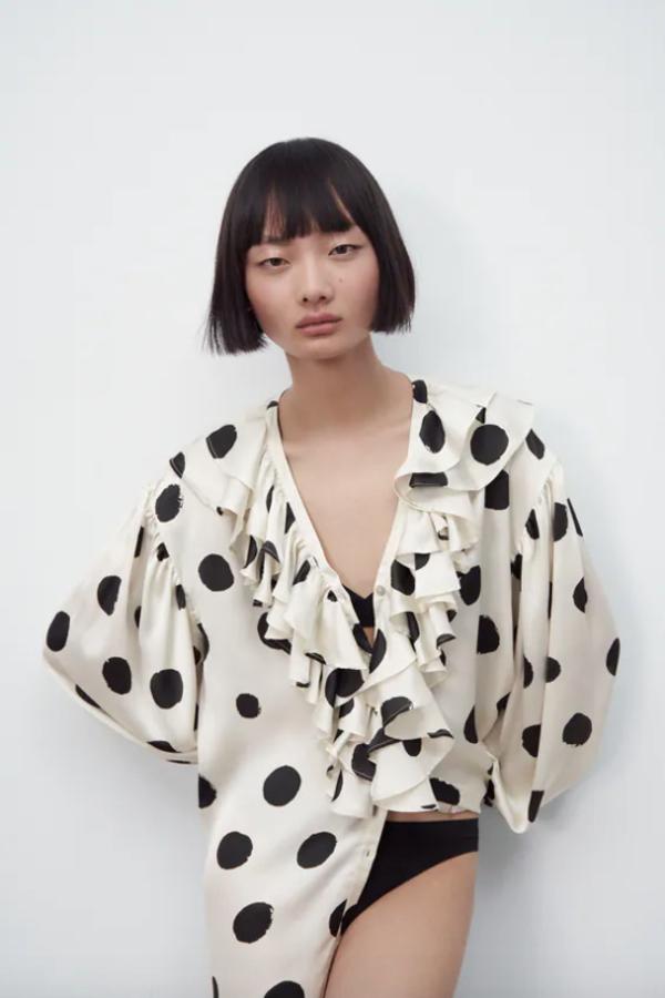 Escote de pico y mangas largas acabadas en puño para esta camisa en tejido satinado y color crudo con estampado de lunares en negro. Es de Zara, viene con volantes en el frontal en el mismo tejido y tiene un precio de 29,95 euros.