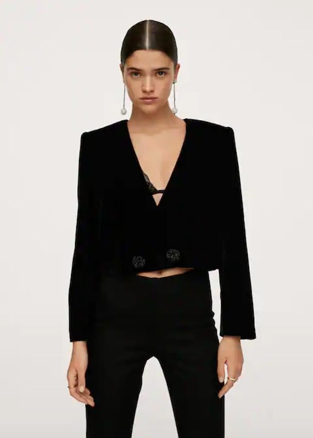 En tejido de terciopelo en color negro, de diseño cruzado recto y con escote de pico, esta blazer cropped de Mango viene con hombreras y cierre de botones frontal en efecto joya. Solo queda en la talla XL y de 49,99 euros ha pasado a costar 29,99 euros. 