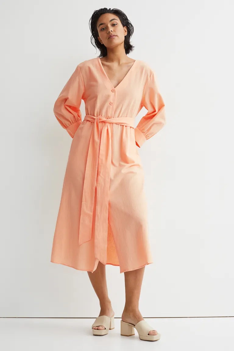 La firma sueca ha apostado por el tono albaricoque para este precioso vestido midi confeccionado en algodón, con escote de pico y botonadura frontal a tono. Con diseño de hombros ligeramente caídos, mangas largas con puños anchos y costura elástica con cinturón de anudar en la cintura, cuesta 39,99 euros y solo se puede comprar en la tienda online de H&M entre las tallas XS y XXL. 