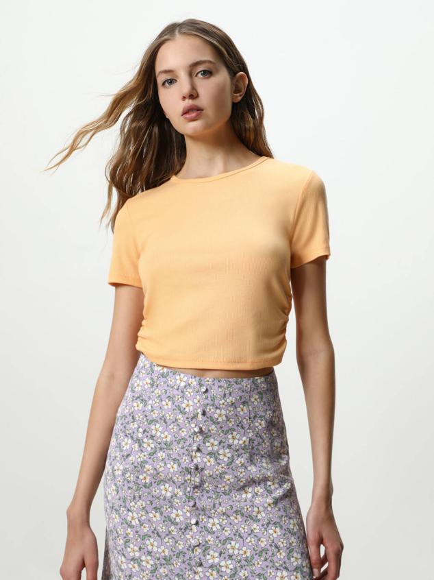 Camiseta en tejido de canalé con manga corta, cuello redondo y detalles fruncidos en los laterales. Está disponible en blanco, naranja, azul claro y negro, con un precio de 5,99 euros.