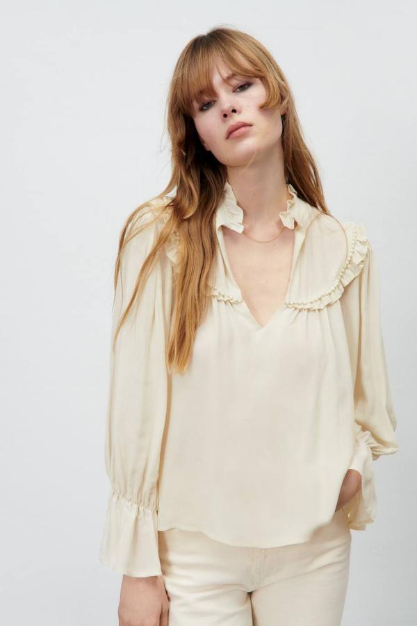 Blusa en tejido fluido y ligero con escote de pico, manga larga, terminaciones de volantes y detalle de pechera con aplicación de perlas a tono, en crudo, de Zara.