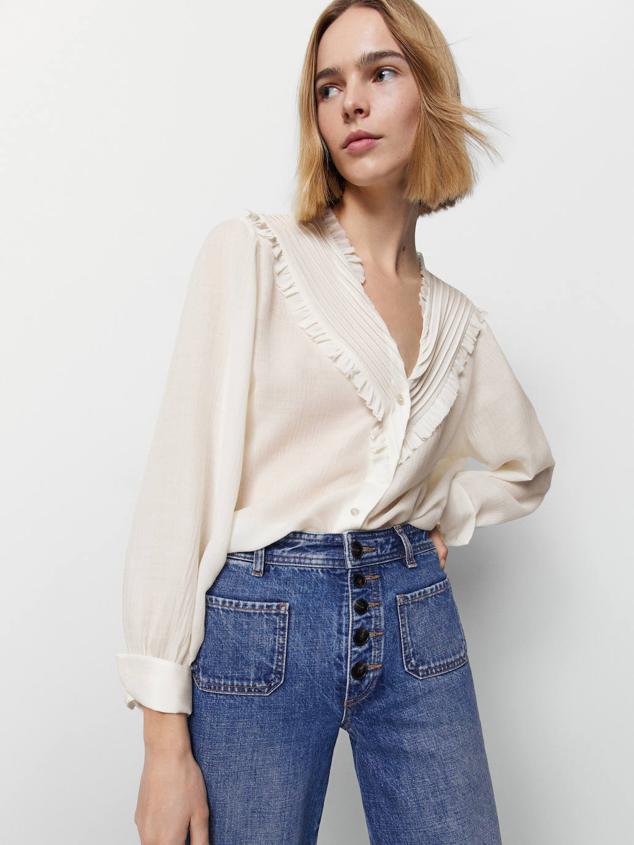 Camisa de inspiración romántica en seda con cuello de pico, adornos de volantes y pechera con detalles plisados, cierre de botones frontal y manga larga recogida en puño, tono crudo. Cuesta 29,95 euros, con descuento.