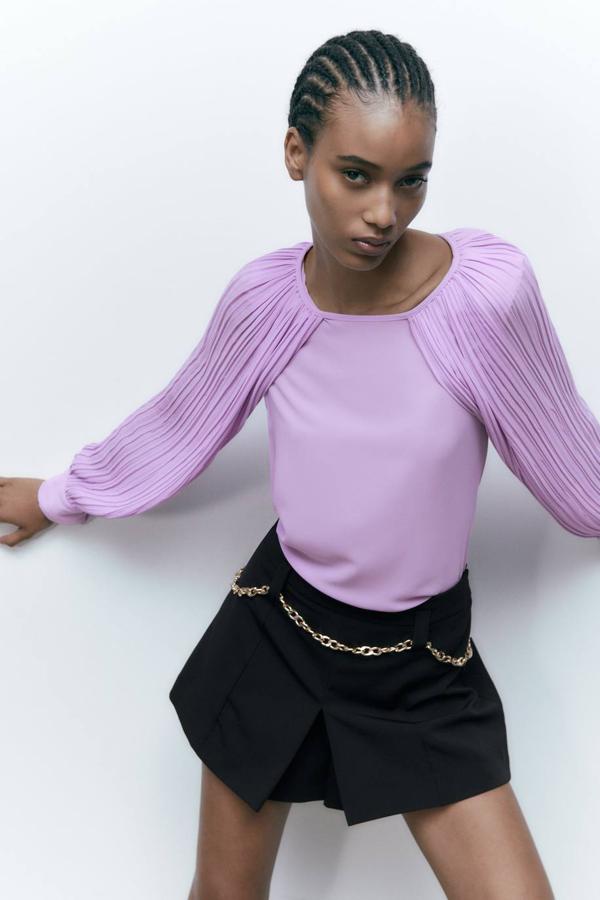 Blusa en tejido plisado con manga larga tipo capa con abertura y cuello redondo, color lila, de Zara. Tiene un precio de 25,95 euros.