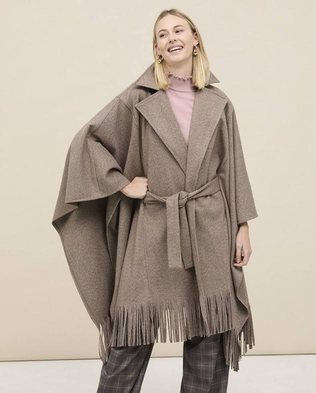 Capa de paño con diseño cruzado, cuello solapa, silueta relajada, mangas amplias, terminaciones de flecos y cinturón de lazada a juego, en marrón, de NKN Nekane. Está disponible a 119 euros. Cómpralo aquí