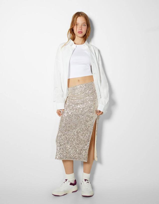Falda midi en tejido elástico con aplicación de lentejuelas, diseño entallado, tiro alto y abertura en el bajo, en champagne, de Bershka. Puedes encontrarla por 29,99 euros.
