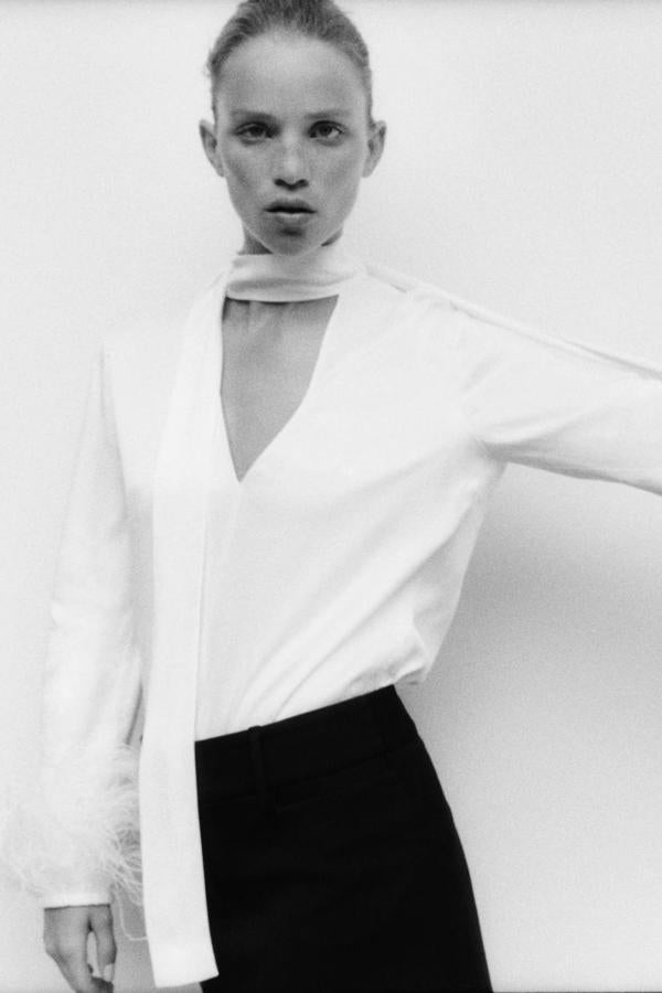 Blusa satinada con corte fluido, escote de pico, lazada al cuello, manga larga y puños de plumas, en blanco, de Zara. Está disponible por 69,95 euros.