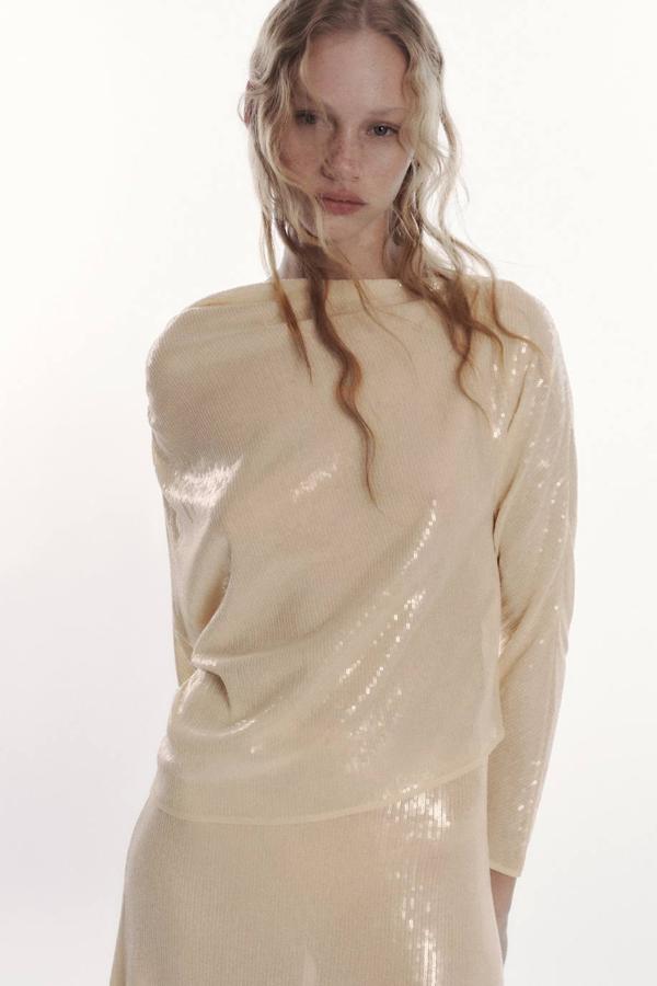 Top confeccionado en tejido de lentejuelas con diseño asimétrico y manga larga, tono crudo, de Zara. Puedes encontrarlo por 39,95 euros.