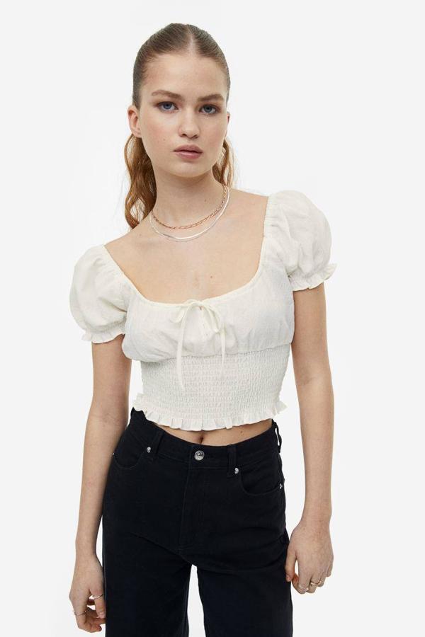 Blusa con escote redondo pronunciado, elástico tipo nido de abeja, lazada y manga corta abullonada con volantes, tono crema, de H&M. Consíguela por 17,99 euros.