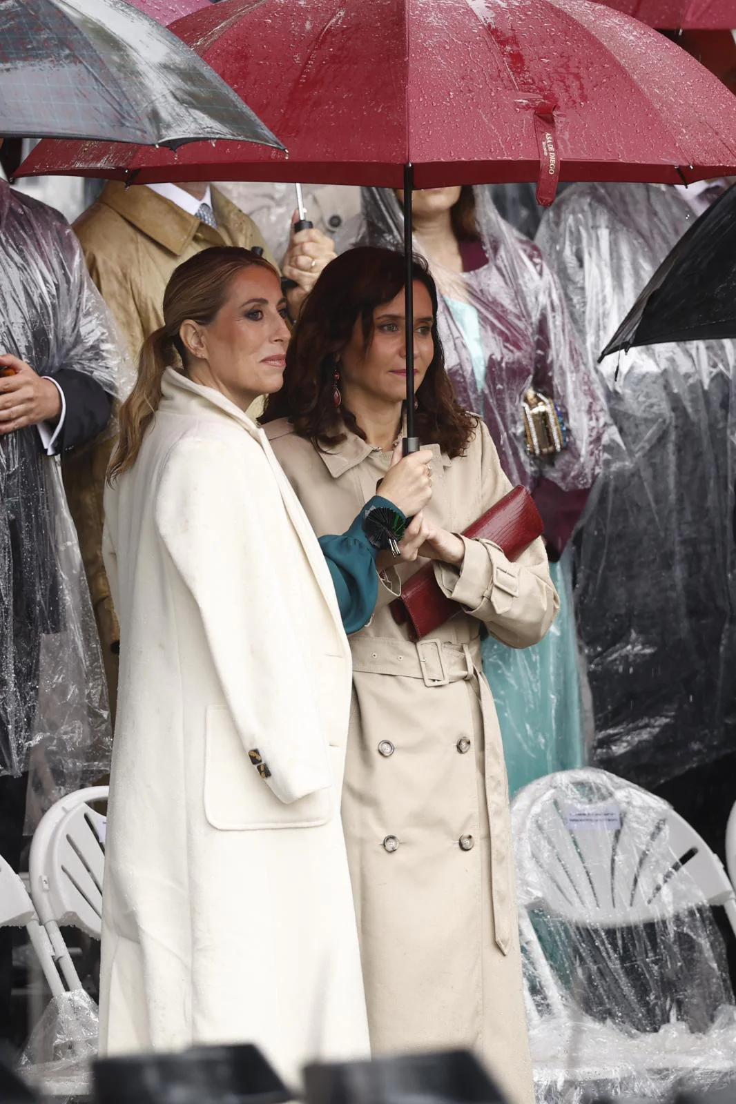 La presidenta de la Comunidad de Madrid, Isabel Díaz Ayuso, y la presidenta de la Junta de Extremadura, María Guardiola, se protegen de la lluvia durante el desfile del Día de la Fiesta Nacional.