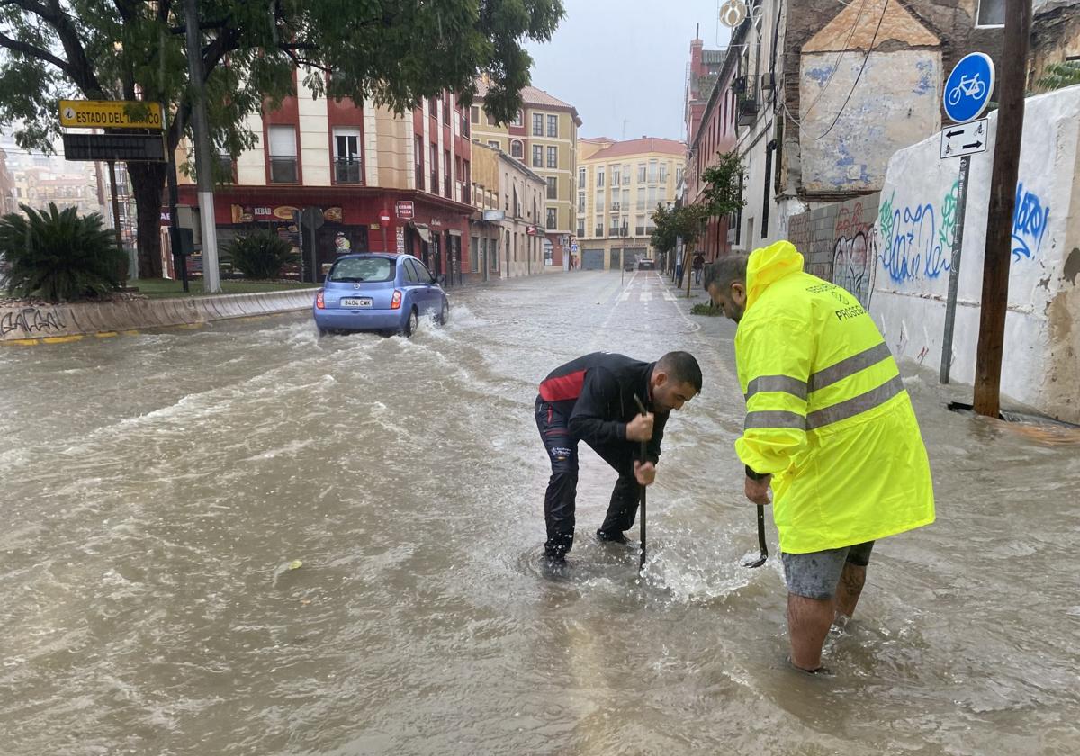 Dos operarios tratan de levantar la tapa de una alcantarilla en una calle inundada de Málaga.