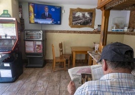 Un vecino de la zona ojea el perfil de Leire Díez en el periódico mientras Núñez Feijóo denuncia el caso en televisión.