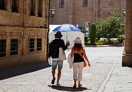 Dos turistas pasean hacia la catedral de Salamanca protegidos con sombreros y paraguas por el sol.