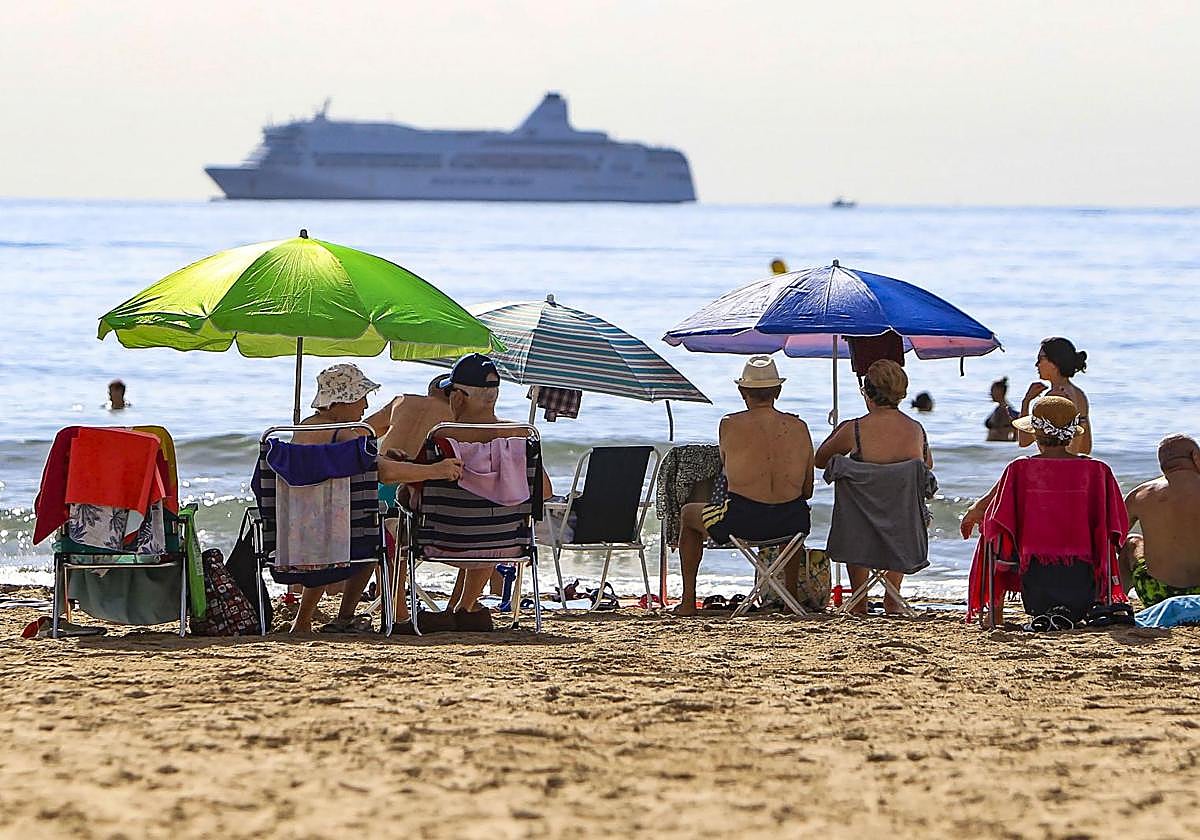 Turistas en la playa del Postiguet ven pasar un crucero.